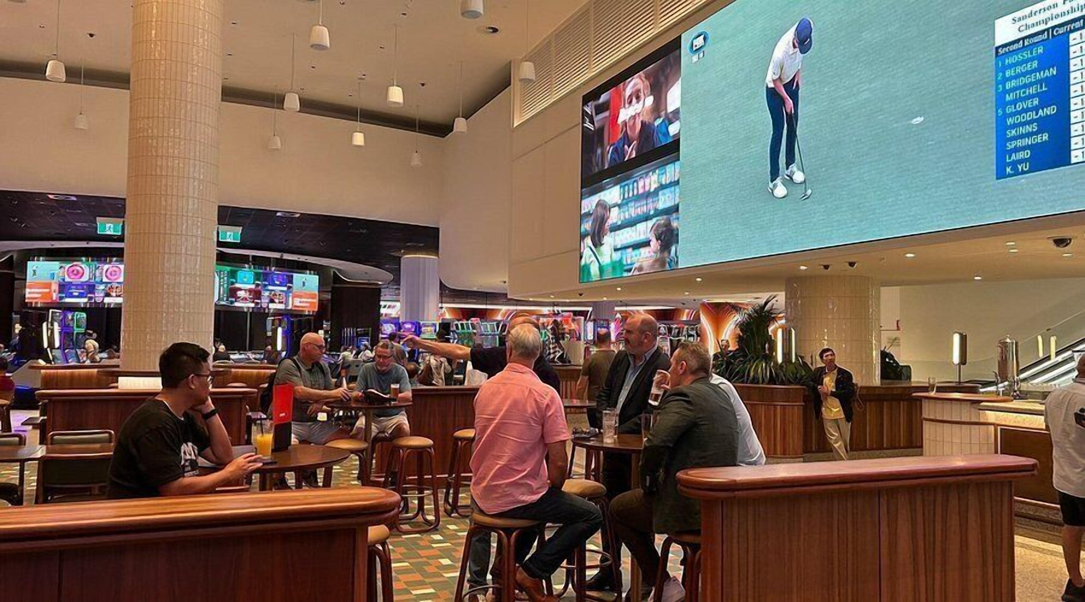 Um espaço amplo e interno com diversas opções de assento, uma tela grande exibindo conteúdo esportivo e um bar.