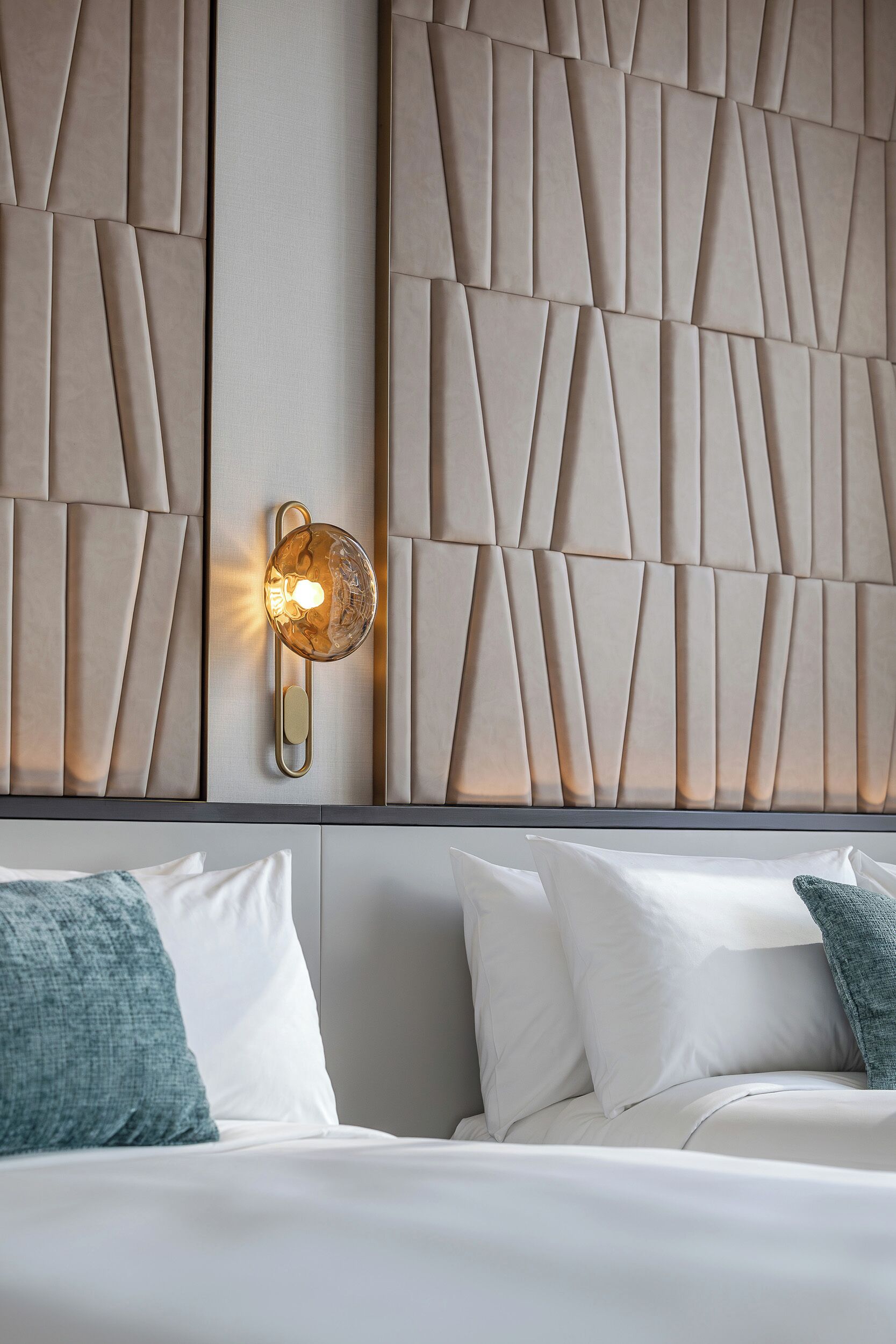 A hotel room with a modern headboard, a wall-mounted light fixture, and neatly arranged pillows.