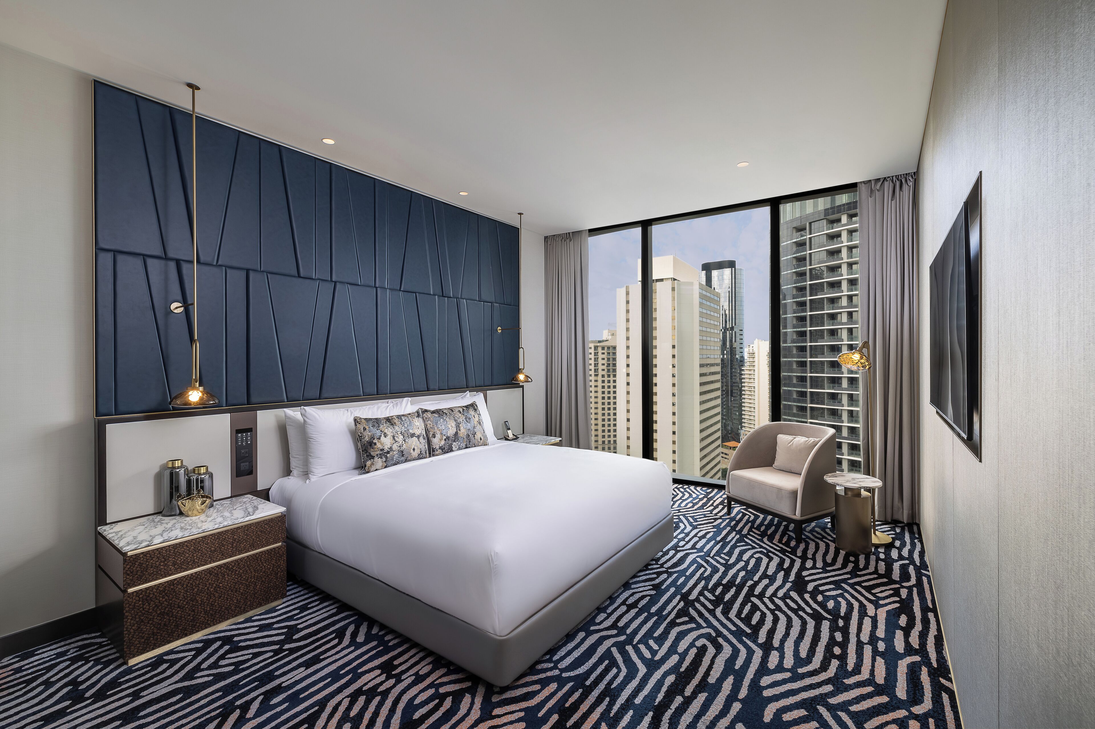Ein modernes Hotelzimmer mit einem großen Bett, einem Nachttisch, einem Sessel und Blick auf hohe Gebäude.