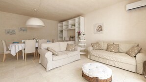 A living room with two sofas, a dining table, chairs, a wooden ottoman, a white shelf unit, and a hanging light fixture. - Villa Reginella - Reginella Deluxe (Lecce)