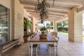 An outdoor patio with a wooden table and white chairs, surrounded by potted plants and a hanging light fixture.