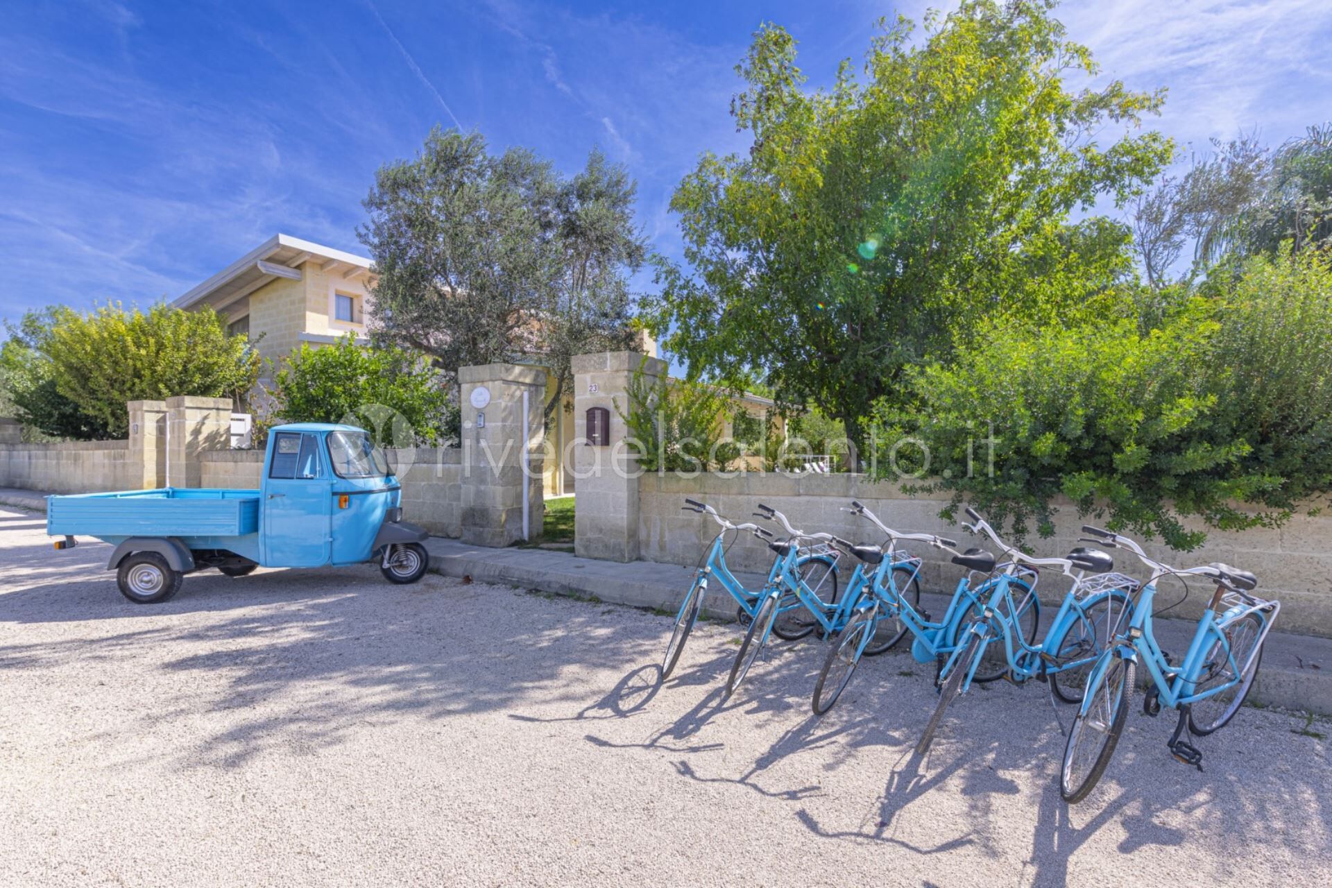 Un camioncino blu parcheggiato su una strada sterrata, con una fila di biciclette blu appoggiate a un muro.