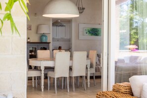 Un coin repas avec une table et des chaises blanches, un luminaire suspendu et une vue sur l’extérieur grâce à une porte vitrée.