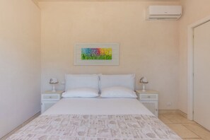 A bedroom with a bed, two bedside tables, a wall-mounted air conditioner, and a colorful painting above the bed.
