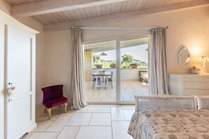 Ein Schlafzimmer mit Bett, Sessel, Kommode und Blick auf eine Terrasse mit Tisch und Stühlen.