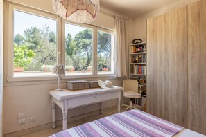A bedroom with a large window, a bedside table with a lamp, a chair, a bookshelf, and a bed with a striped bedspread.
