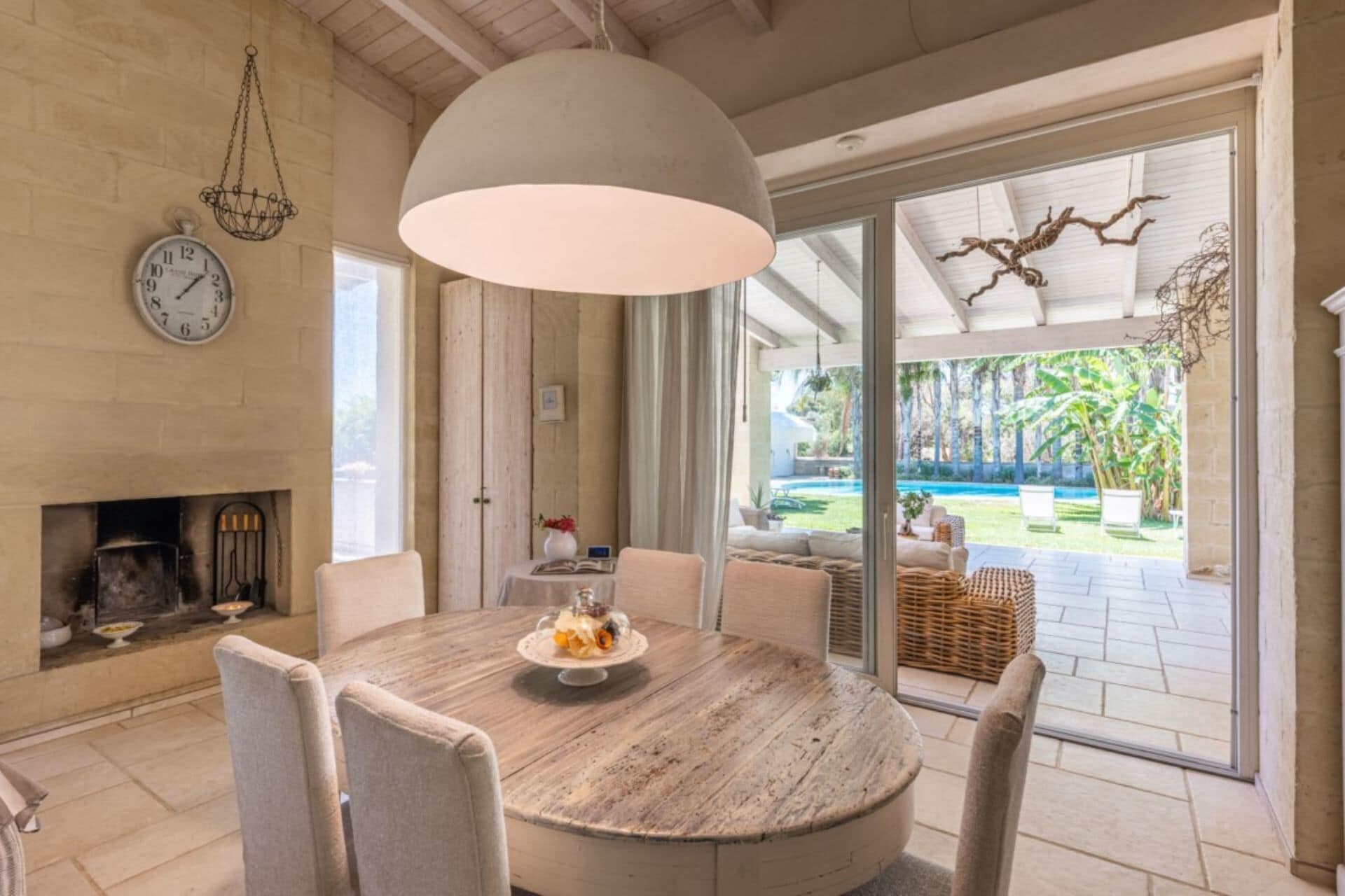 Un coin repas avec une table ronde, des chaises, un luminaire suspendu, une horloge et une vue sur une piscine et un jardin.