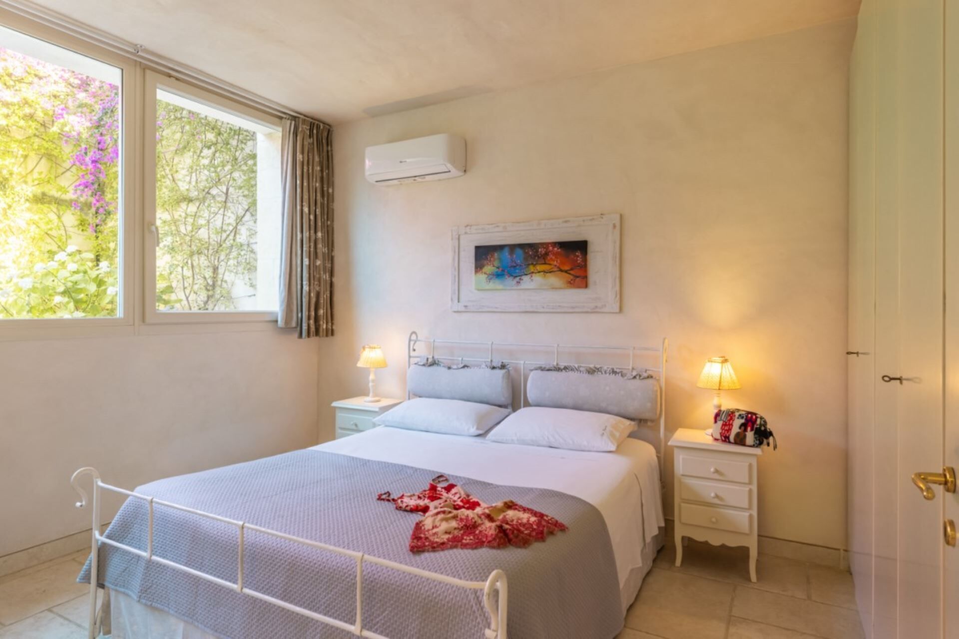 Una camera da letto con un letto, due comodini, una finestra con tende, un condizionatore d'aria e un quadro appeso al muro.
