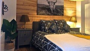A bedroom with a wooden wall, a bed with patterned bedding, a nightstand with a lamp, and a painting of a bull.