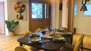 A dining area with a dark wooden table set for four, surrounded by yellow chairs. A potted plant is on the left, and a colorful abstract painting is on the wall.