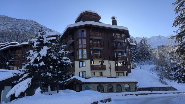 A multi-story lodge with balconies and a stone facade, surrounded by snow-covered trees and mountains.