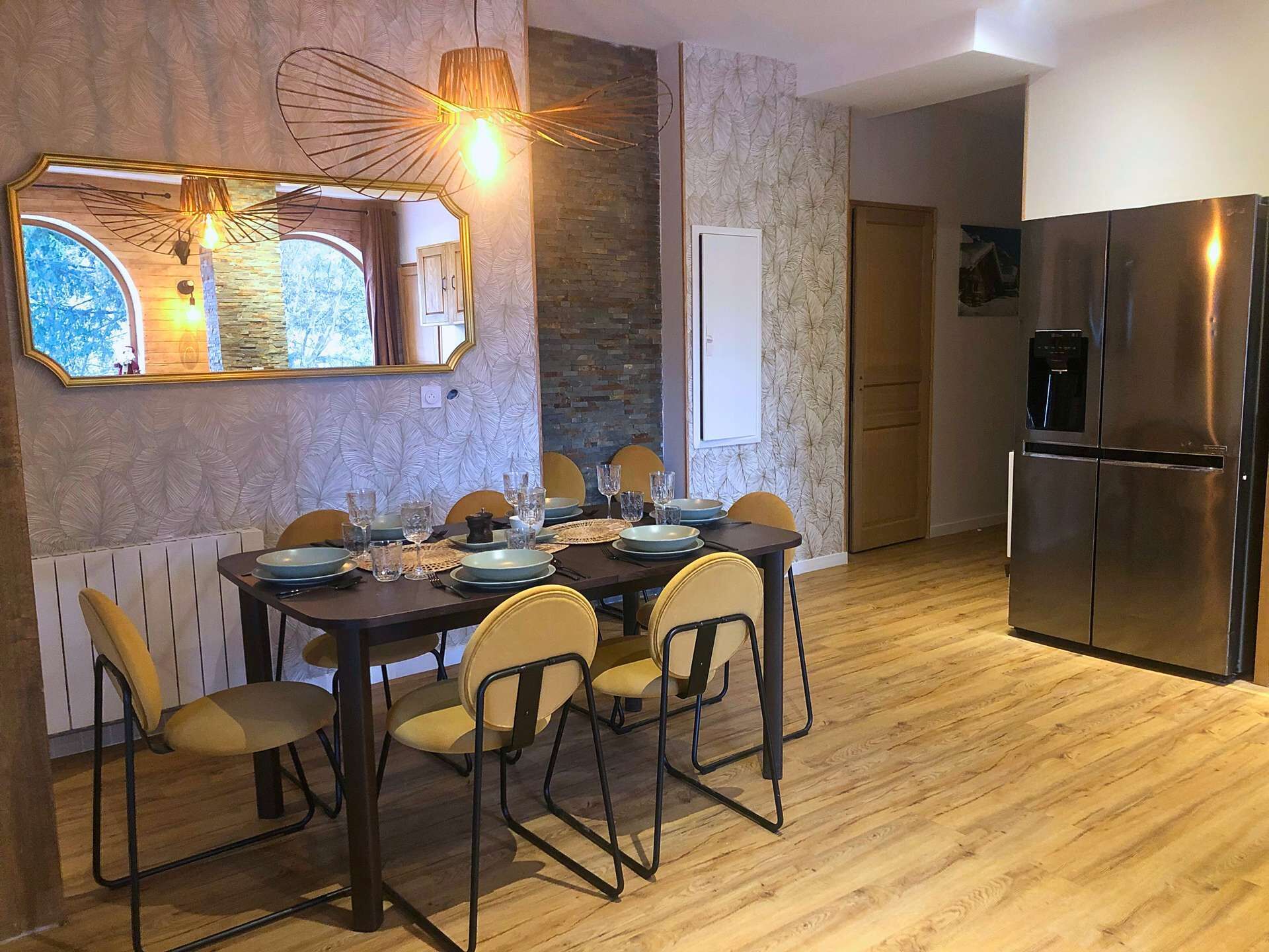 Un coin repas avec une table en bois dressée pour six personnes, entourée de chaises. Un grand miroir est accroché au-dessus de la table et reflète la pièce. On aperçoit un réfrigérateur moderne à l’arrière-plan.