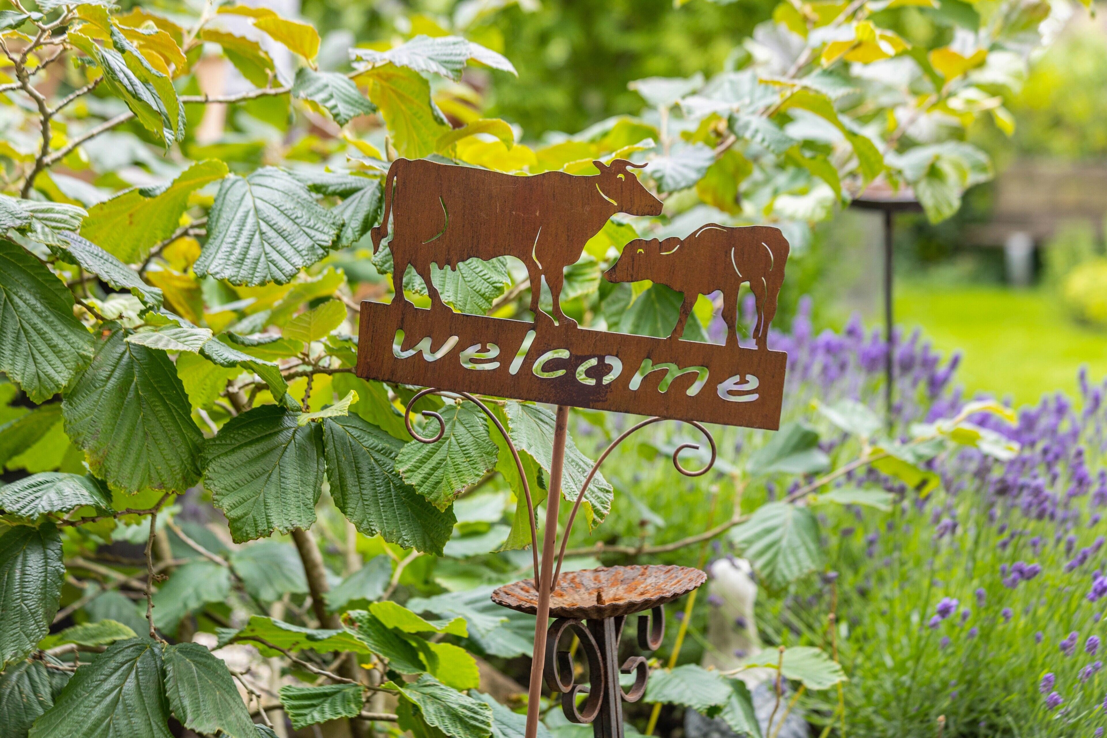 A metal sign with a cow silhouette and the word 'welcome' in a garden setting.