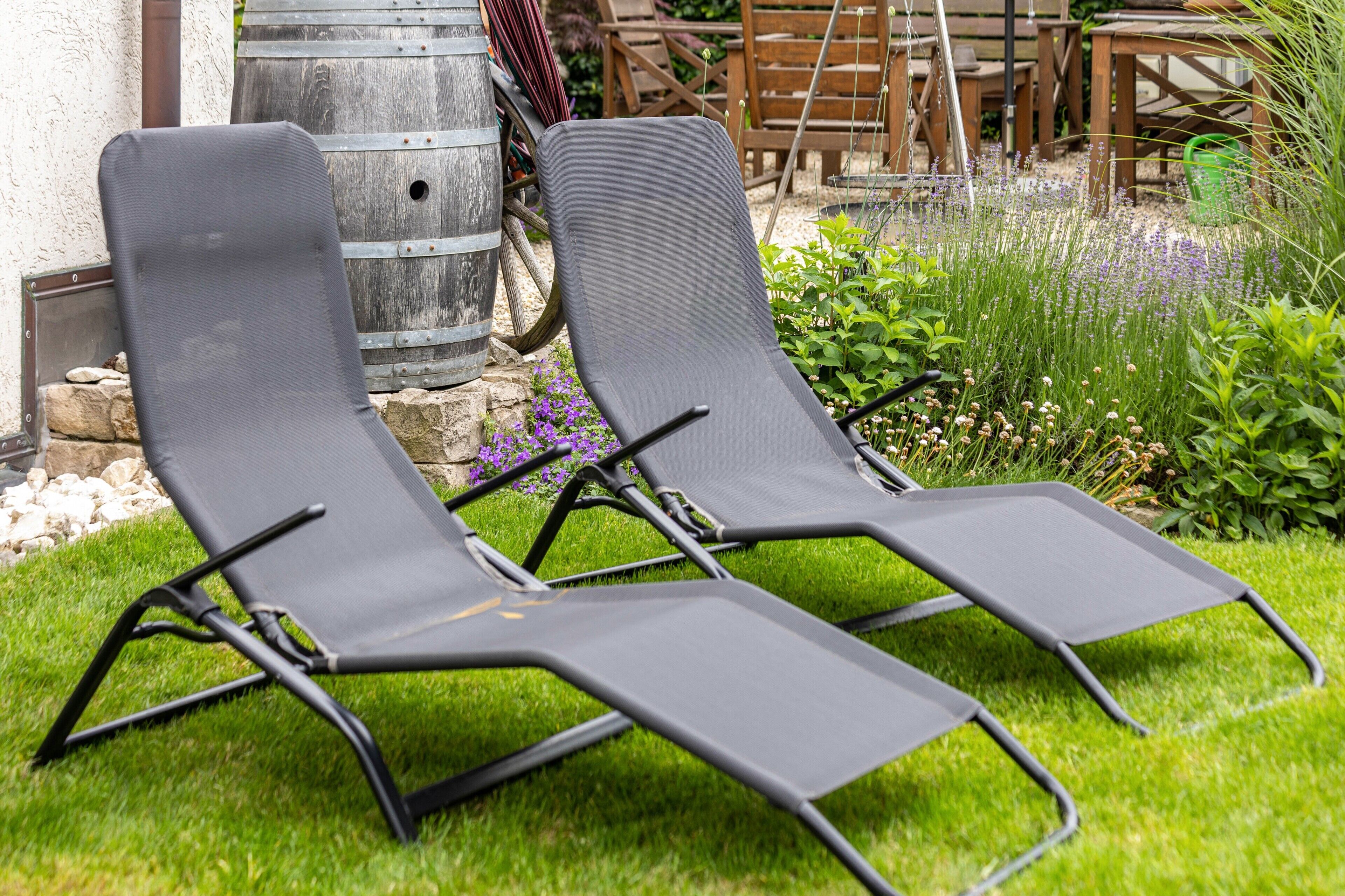Zwei schwarze Lounge-Stühle mit Metallgestell, auf einer Rasenfläche mit einer Steinmauer und Holzmöbeln im Hintergrund.