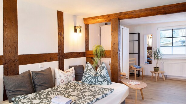 Ein Zimmer mit einem Bett, Holzbalken, einem kleinen Tisch und einer Topfpflanze.