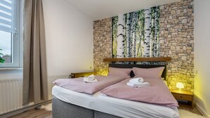 A double bed with checkered bedding, a stone wall with birch tree artwork, a window with curtains, and a bedside table with a lamp.