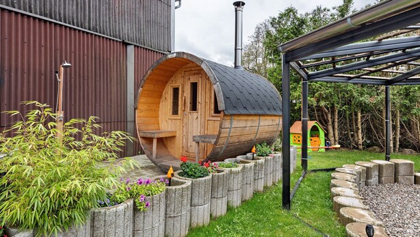 A wooden barrel-shaped structure with a door, a metal roof, and a garden with plants and a small wooden playhouse in the background.