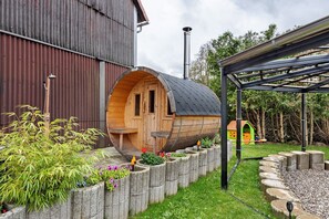 Una estructura cilíndrica de madera con puerta, techo de metal y un jardín con plantas y una pequeña casita de madera al fondo.