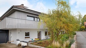 Une maison moderne à deux étages avec un garage, située au numéro 24, et un jardin agrémenté de diverses plantes et d’un arbre.