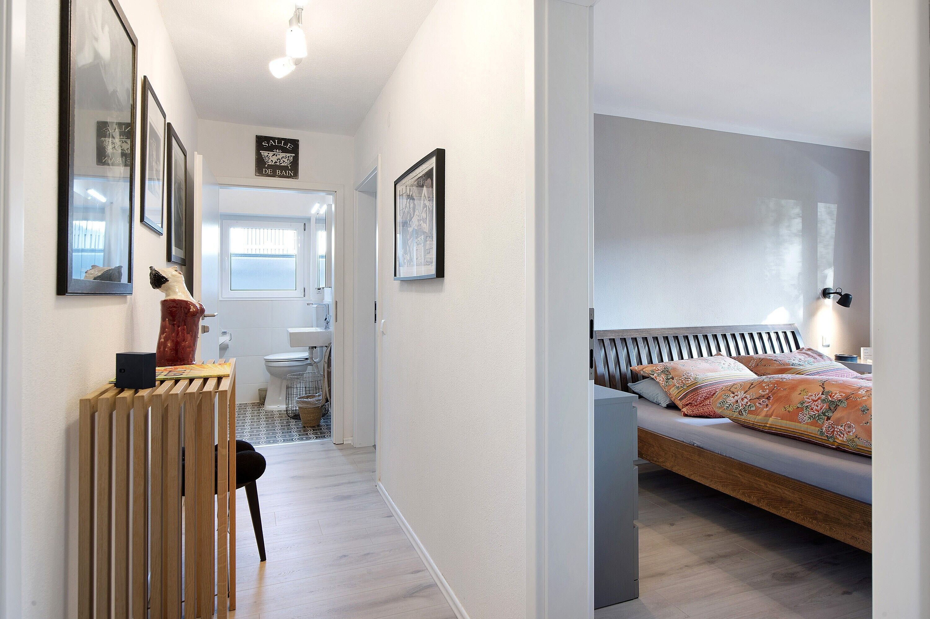 A bedroom with a bed, a bedside table, and a chair. There is a bathroom with a toilet and sink. The room has white walls and a wooden floor.