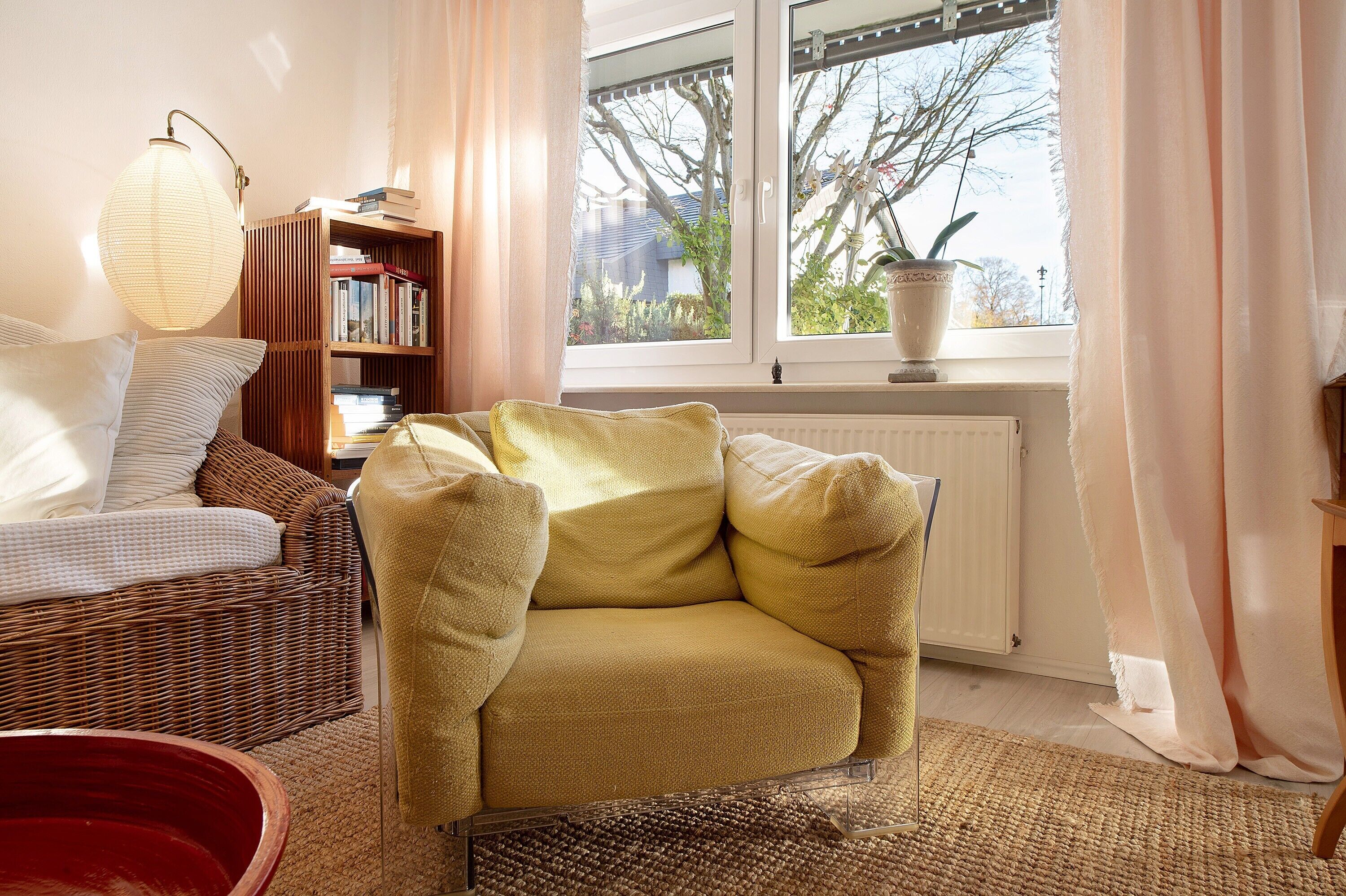 Un salon chaleureux avec un canapé en osier, un fauteuil jaune, une bibliothèque et une fenêtre donnant sur des arbres et des immeubles.