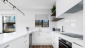 Une cuisine moderne avec des meubles blancs, un four encastré et une fenêtre donnant sur un immeuble.