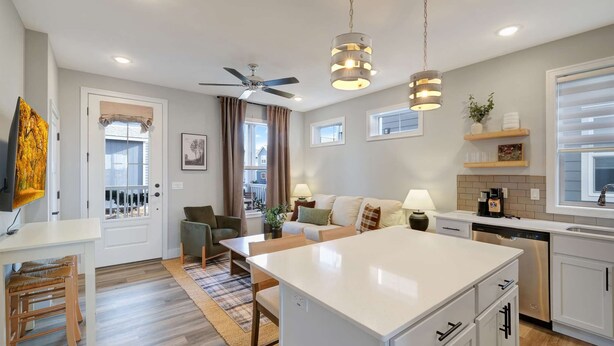 A modern kitchen with a central island, a dining area with a table and chairs, a ceiling fan, pendant lights, and a view of the outside. - Mansion Flat @ Navvy Town (Ottawa)