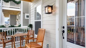 A covered porch with wooden chairs and a table, a white door with a window, and a wall-mounted lantern. - Mansion Flat @ Navvy Town (Ottawa)