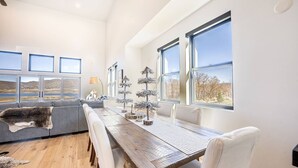 Ein geräumiger Wohnbereich mit Esstisch aus Holz, grauem Sofa und großen Fenstern mit reizvoller Aussicht.