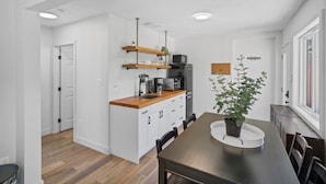 A modern kitchen with white cabinets, a wooden countertop, and a built-in oven. There is a dining area with a table and chairs, and a potted plant on the table. - Spacious Riverside Suite | Kitchenette, BBQ, Dock (Lake Cowichan)