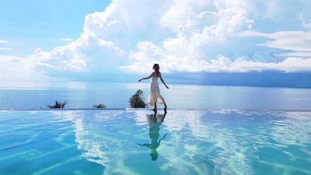 A woman in a white dress standing on a clear blue infinity pool with a view of the ocean and sky. - Apec Mandala  View Bien (Hoa Thang)
