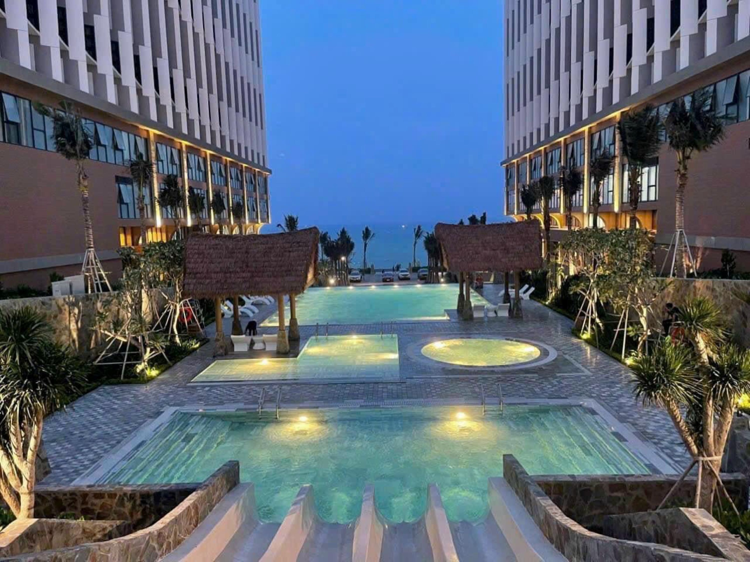 A resort pool area with a central water feature, surrounded by lounge chairs and palm trees.