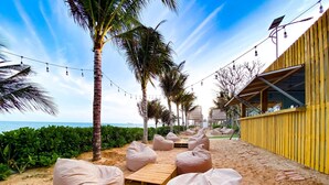 A beachside seating area with bean bags and a wooden table, palm trees, and a yellow building with a sloped roof. - Apec Mandala  View Bien (Hoa Thang)