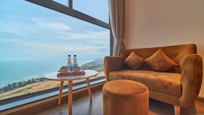 A room with a large window overlooking a beach, a plush armchair, and a small round table with bottles and glasses. - Apec Mandala  View Bien (Hoa Thang)