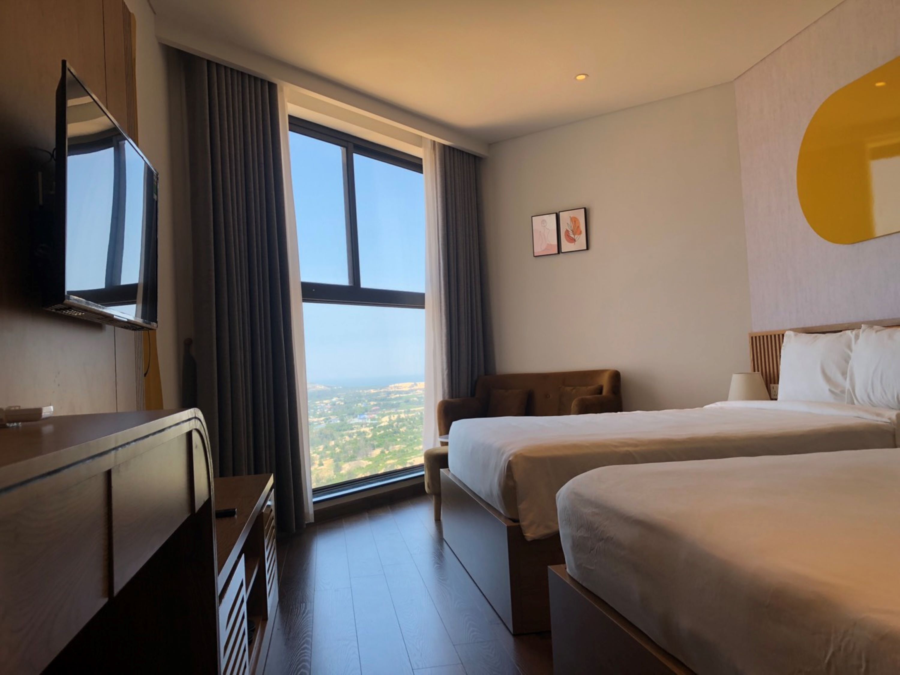 A hotel room with a large window, a flat-screen TV, two beds, and a wooden dresser.