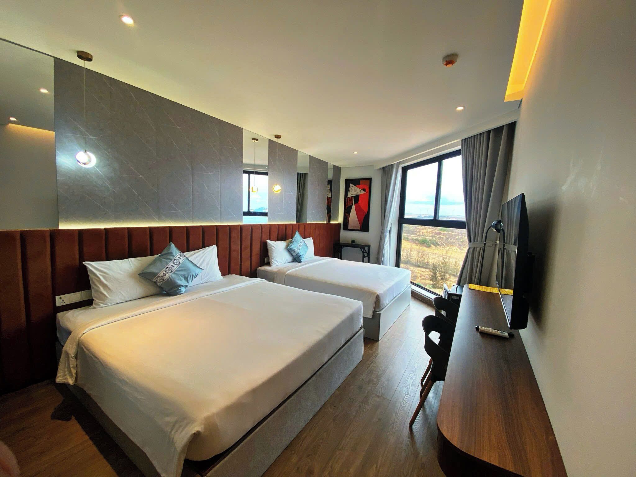 A hotel room with two beds, a wooden desk, a flat-screen TV, and a large window offering a view.