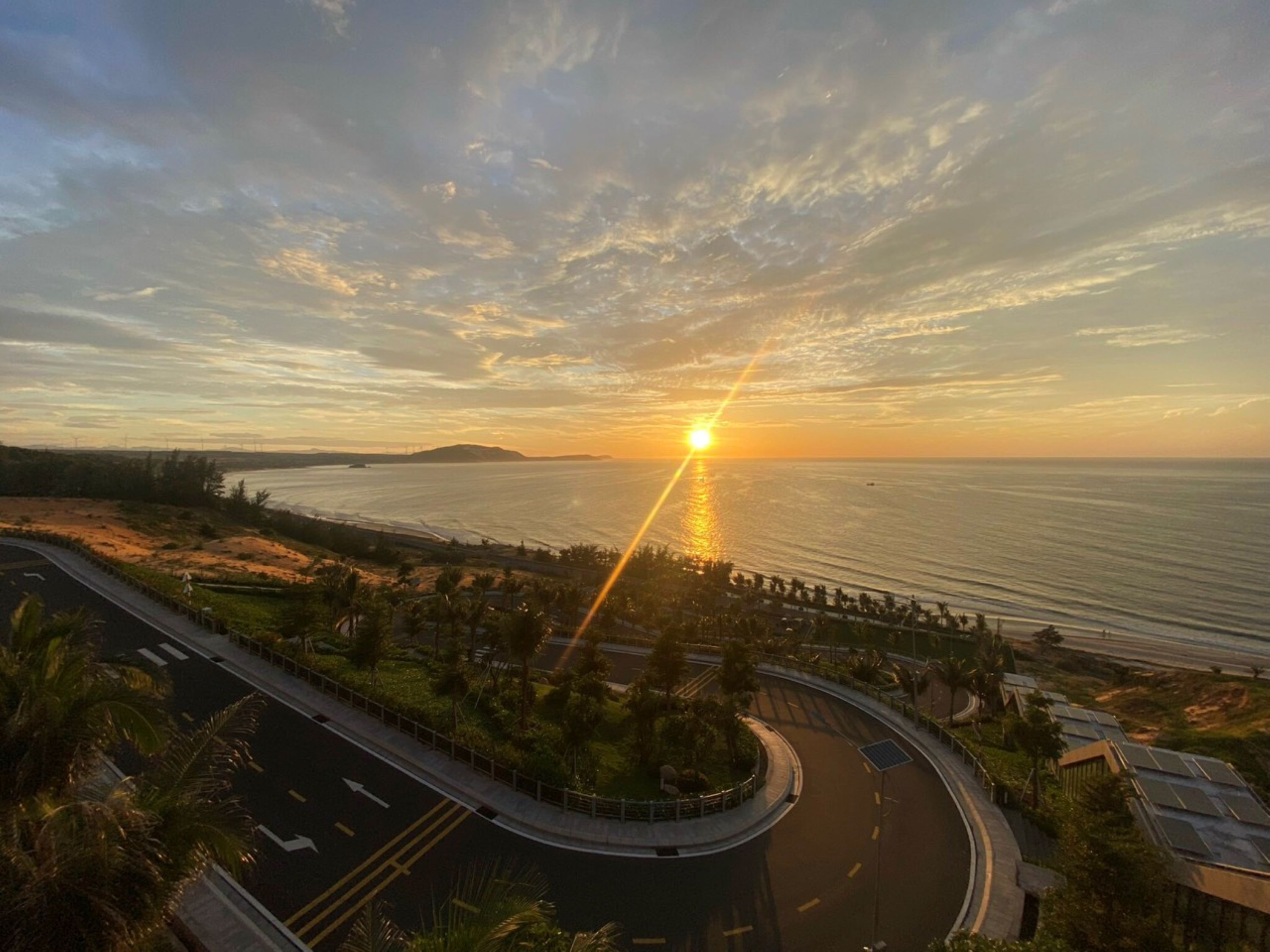 Eine Küstenstraße schlängelt sich zum Meer, im Hintergrund ein Sonnenuntergang.