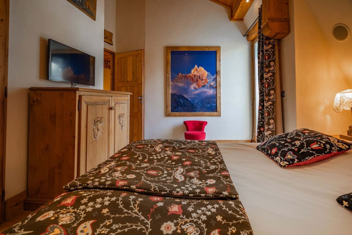 A bedroom with a bed, a wooden wardrobe, a red chair, a painting of a mountain, and a window with curtains.