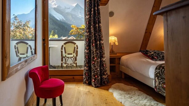 A bedroom with a bed, a red chair, a window with a view of mountains, and a wooden ceiling.