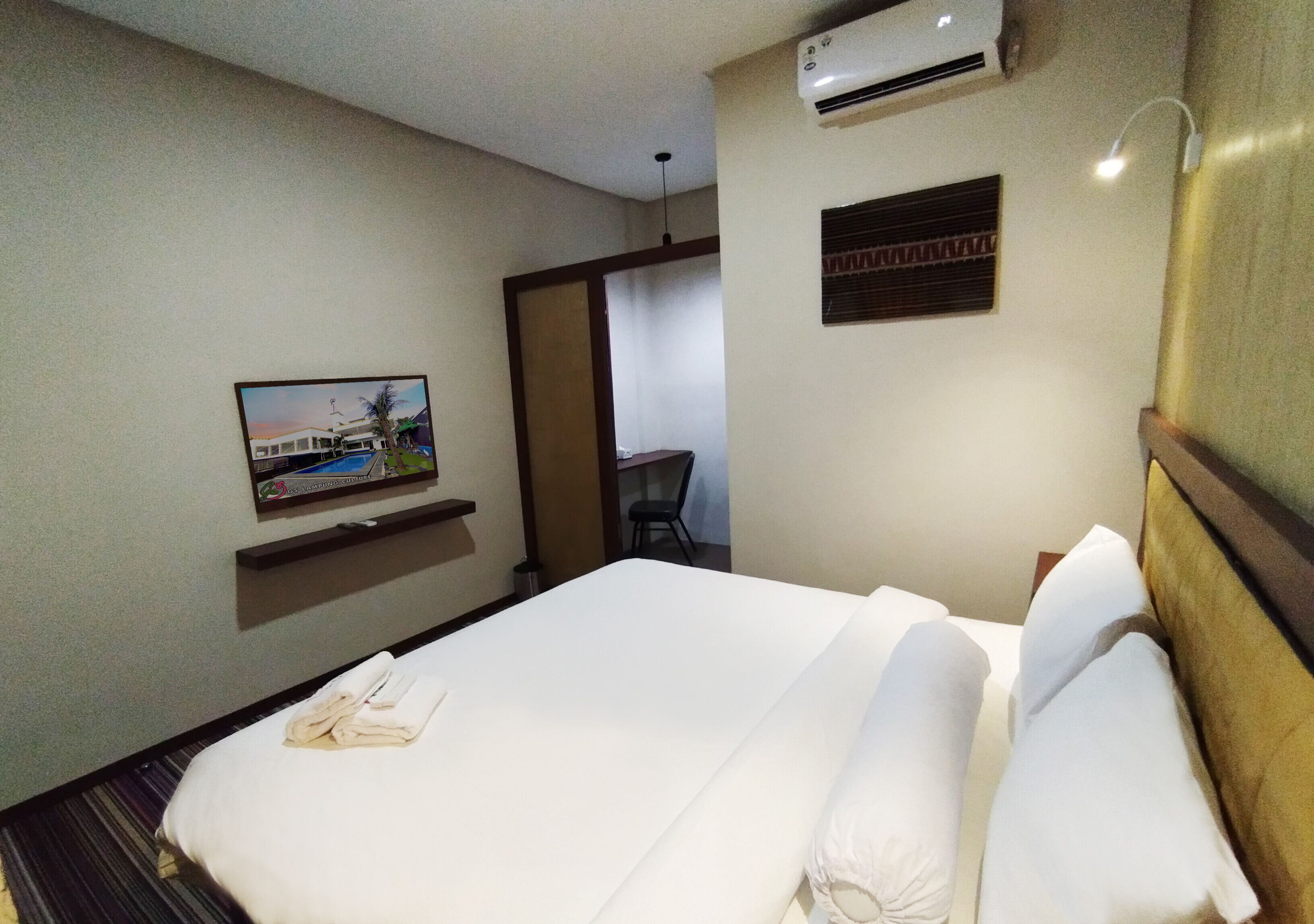 A hotel room with a large bed, a flat-screen TV mounted on the wall, an air conditioning unit, and a small table with a chair in the corner.
