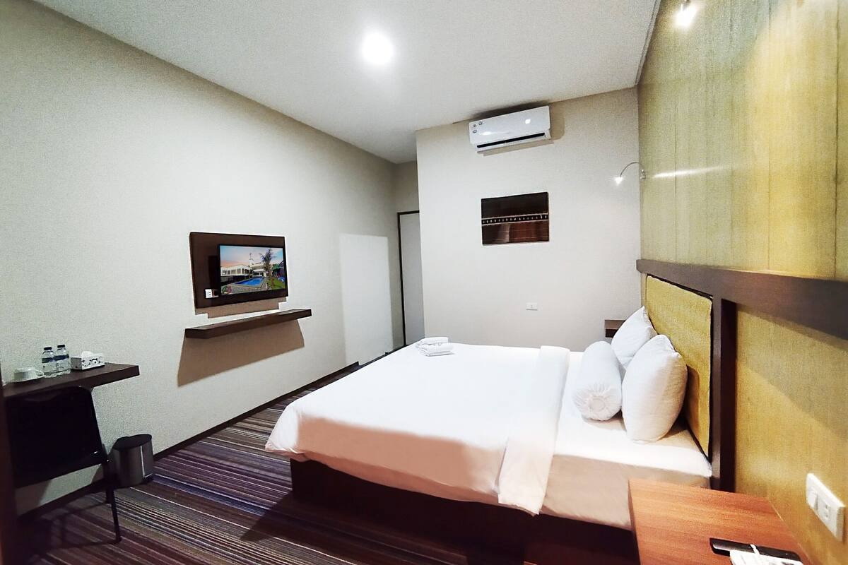 A hotel room with a bed, a desk, a TV mounted on the wall, an air conditioning unit, and a small window.