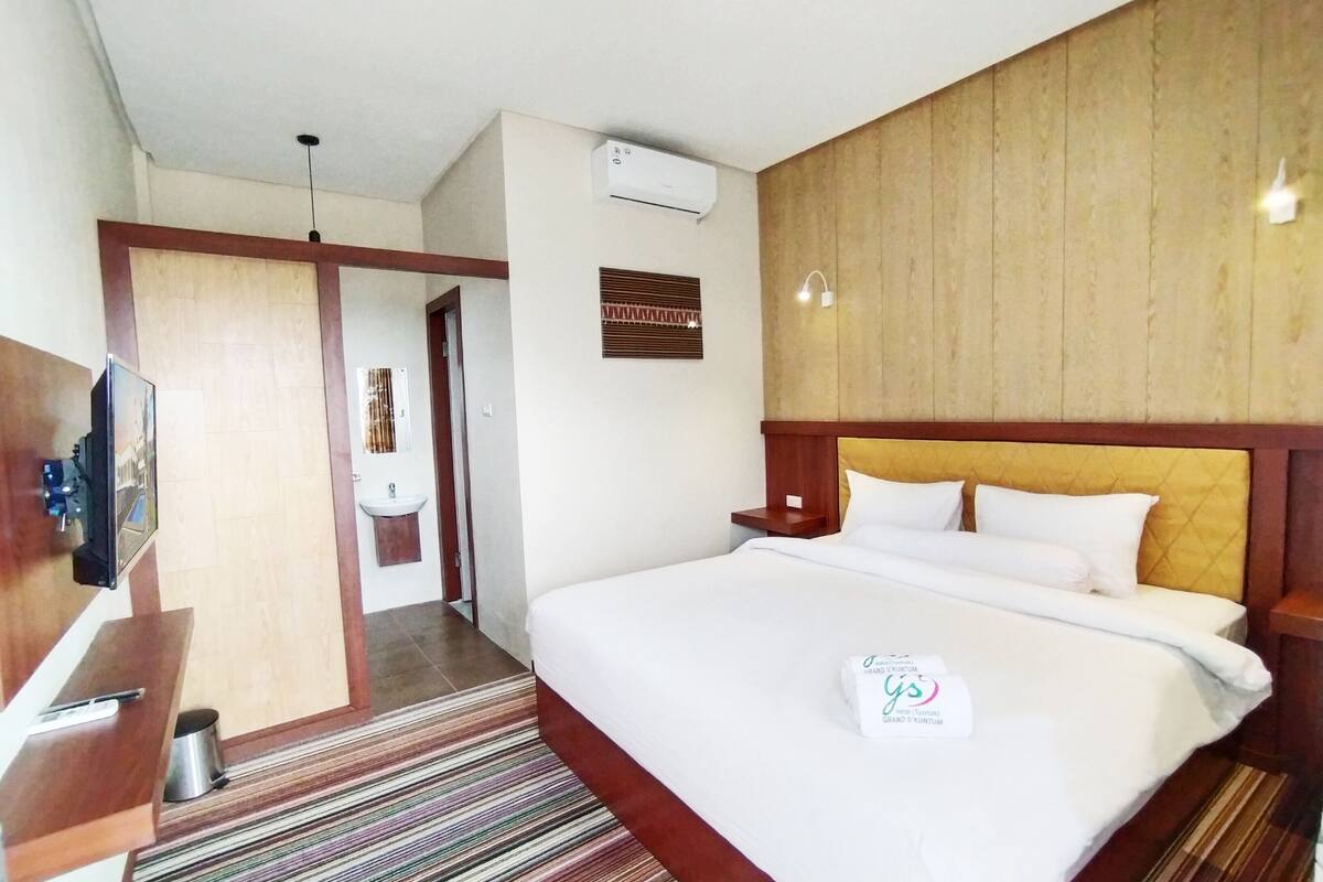 A hotel room with a large bed, a bedside table, a TV, an air conditioning unit, and a wooden headboard.