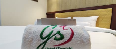 A neatly made bed with a white towel featuring embroidered text, a wooden headboard, and a bedside table with a 'Dental Kit' sign.