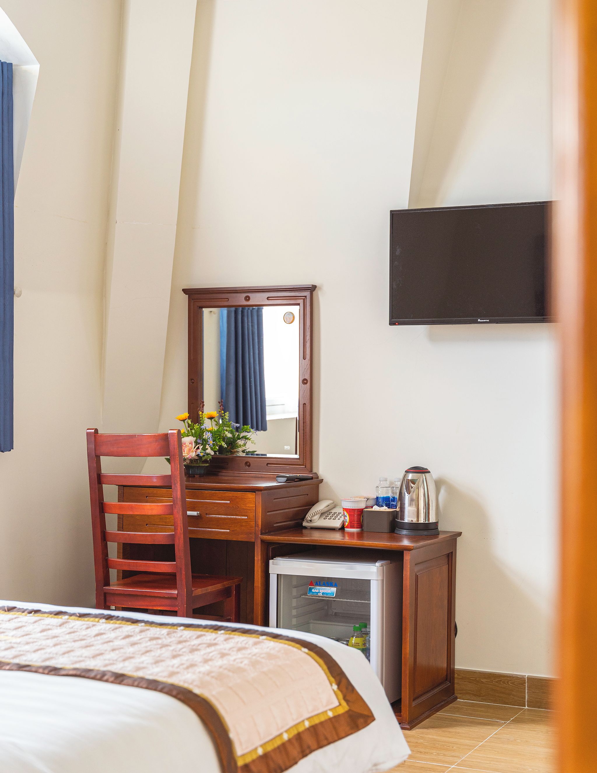 Una habitación de hotel con cama, un escritorio de madera con teléfono, un refrigerador pequeño y un televisor montado en la pared.