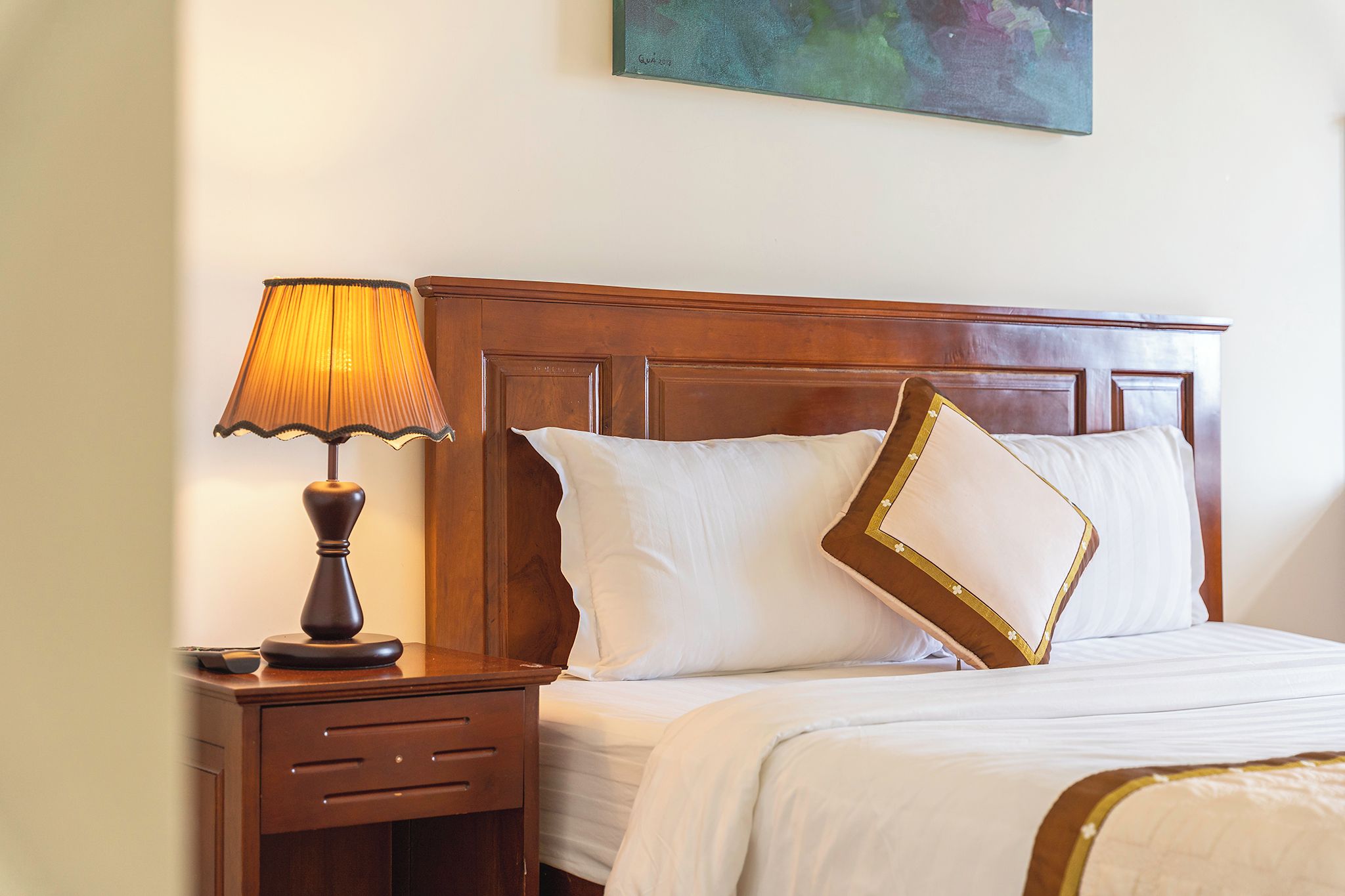 A hotel room with a wooden headboard, a bedside lamp, a nightstand, and a bed with white bedding and decorative pillows.