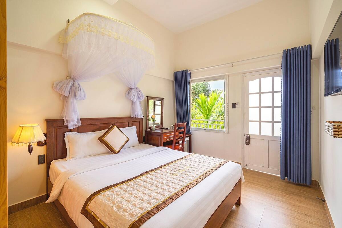 A hotel room with a bed, a nightstand, a lamp, a window with curtains, and a view of greenery outside.