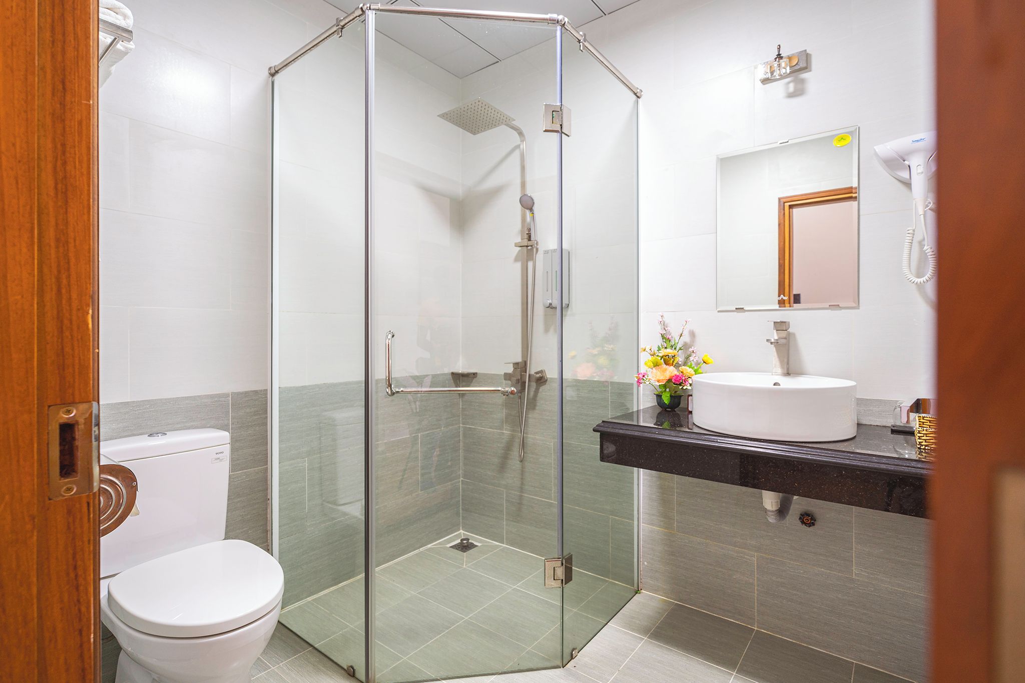 A modern bathroom with a glass-enclosed shower, a white toilet, a sink with a mirror above, and a vase of flowers on the counter.