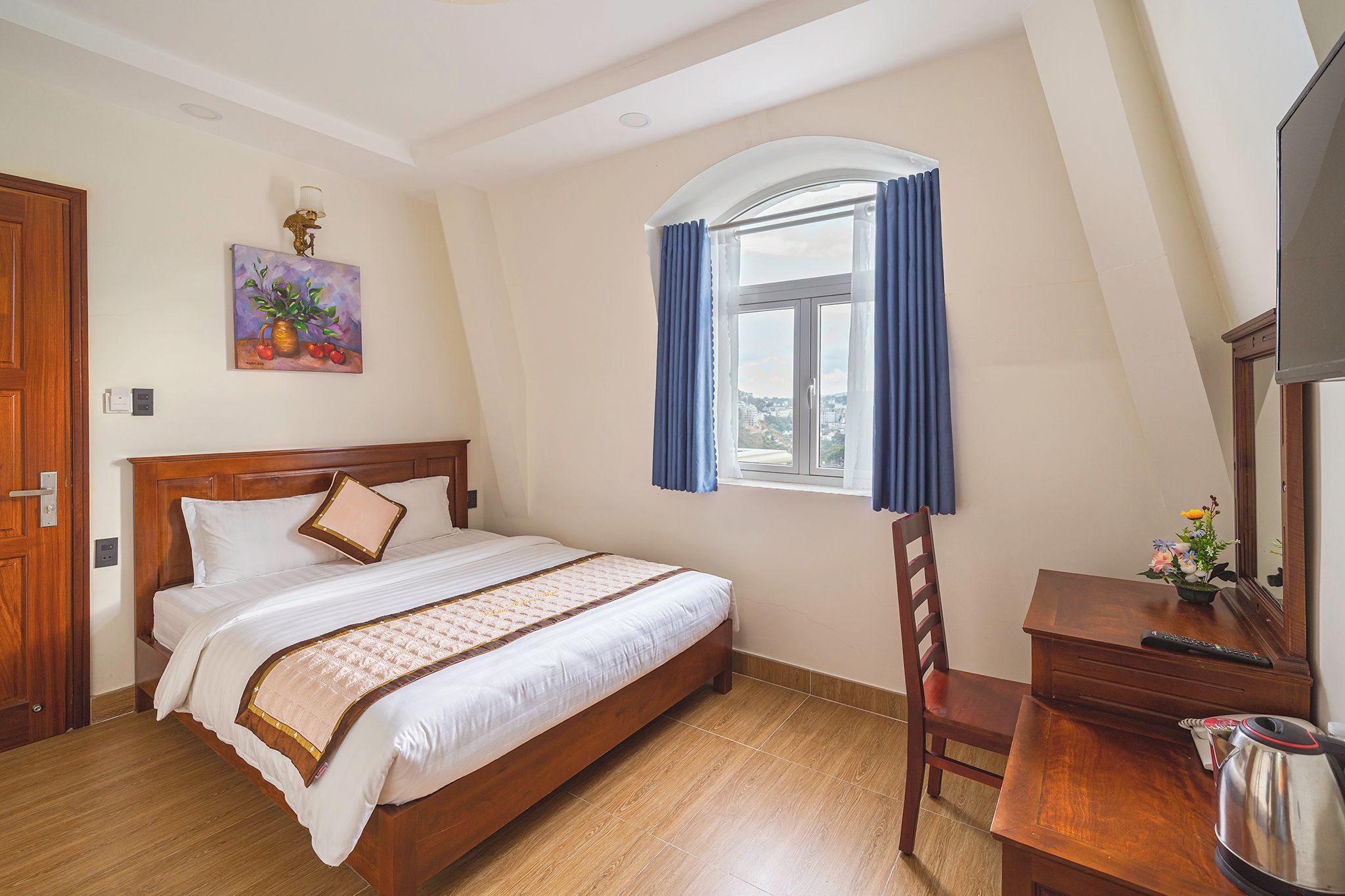 A bedroom with a wooden bed, a desk with a chair, a window with curtains, and a painting on the wall.