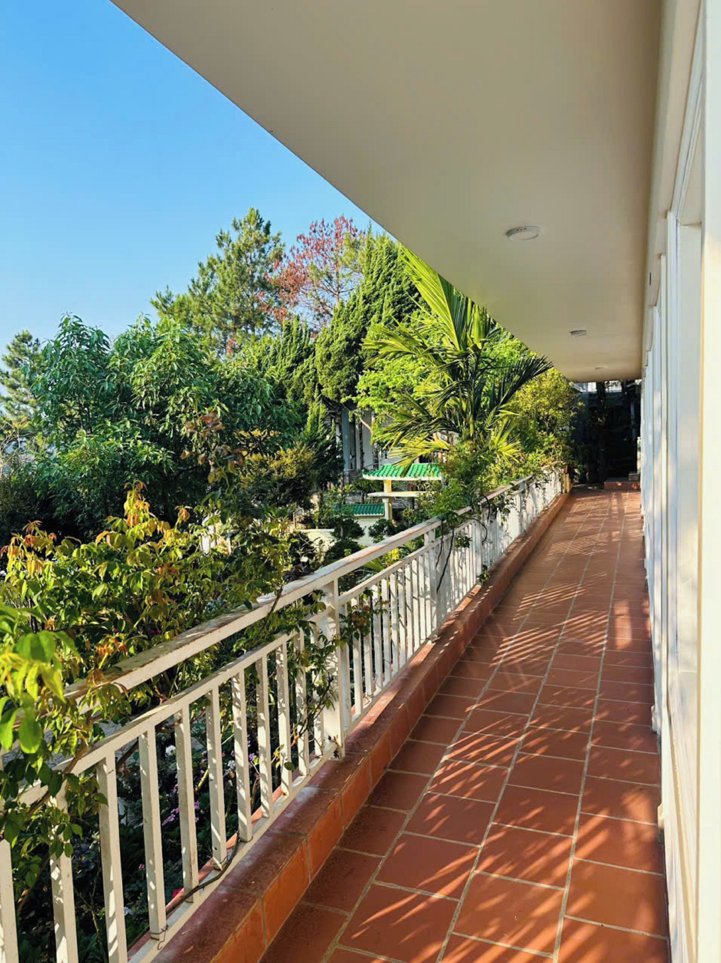 A covered balcony with a tiled floor, surrounded by lush greenery and a white railing.