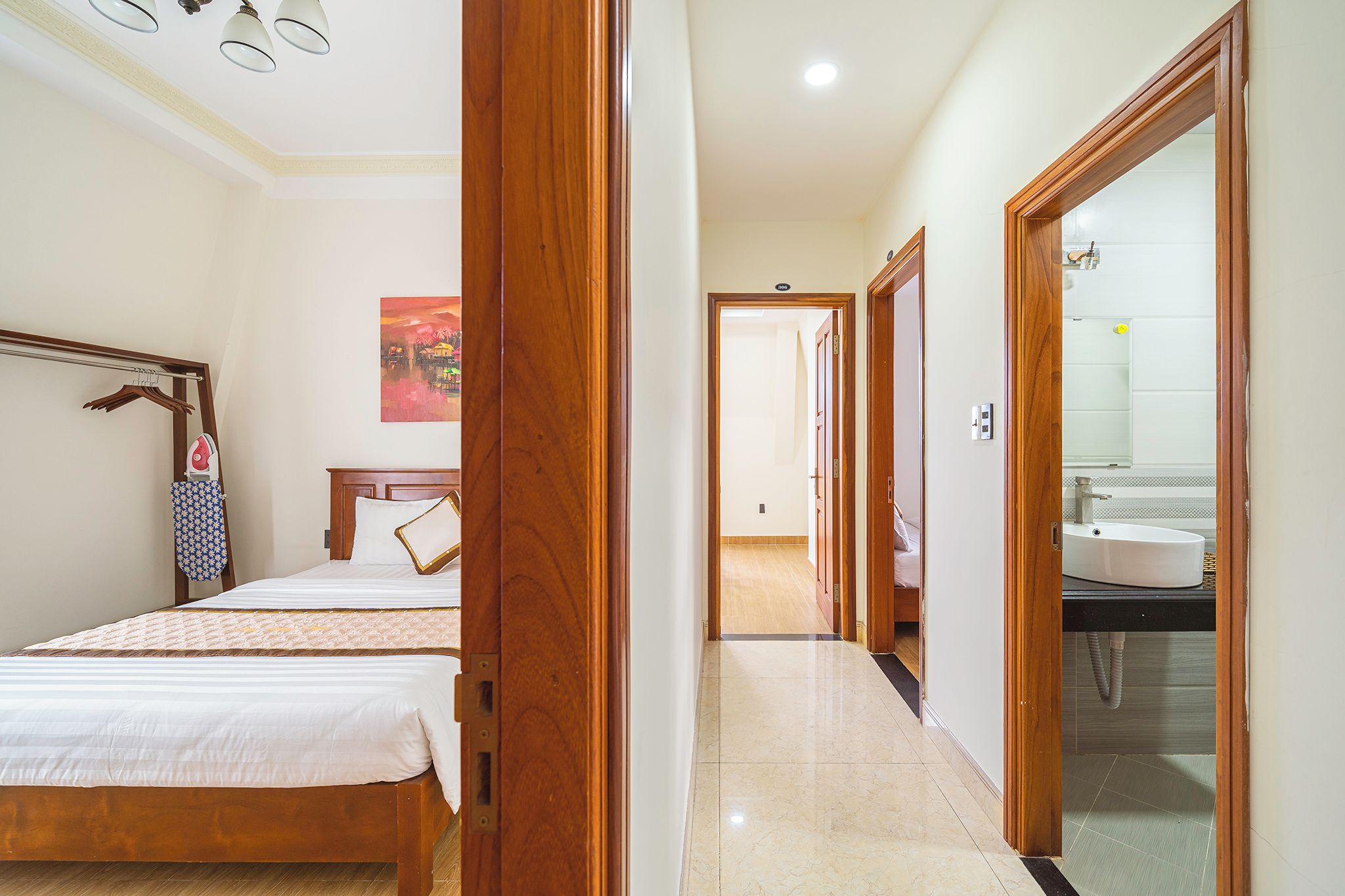 A hotel room with a bed, a wooden wardrobe, a bathroom with a sink, and a hallway leading to another room.